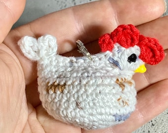 Pode incluir: Um pequeno ornamento de galinha em crochê branco com uma crista vermelha brilhante, bico amarelo e um olho preto. O corpo é feito de fio branco com manchas sutis em marrom e azul, e um pequeno anel de metal prateado está preso no topo.