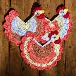 May include: Four handmade crochet chicken-shaped pot holders. Each pot holder features a red comb and wattle, yellow beak, and a body with shades of brown, peach, and blue. The pot holders are arranged on a wooden surface.