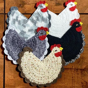 May include: A collection of five handmade crochet chicken-shaped pot holders. The pot holders feature various colors, including white, black, gray, and cream, with red and yellow accents for the comb and wattle. Each has a scalloped edge.