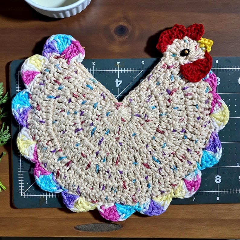 Pode incluir: Um descanso de panela em croch&ecirc; com formato de galinha, feito de fio bege com manchas multicoloridas. A borda apresenta acabamentos festonados em azul, rosa e amarelo. A cabe&ccedil;a tem uma crista e barbelas vermelhas com um bico amarelo.
