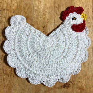 Pode incluir: Um porta-copos ou aplica&ccedil;&atilde;o de croch&ecirc; branco em forma de galinha com borda festonada. O design apresenta uma crista e barbelas vermelho-brilhantes, um pequeno bico amarelo e um olho escuro, tudo feito com pontos de l&atilde; detalhados.