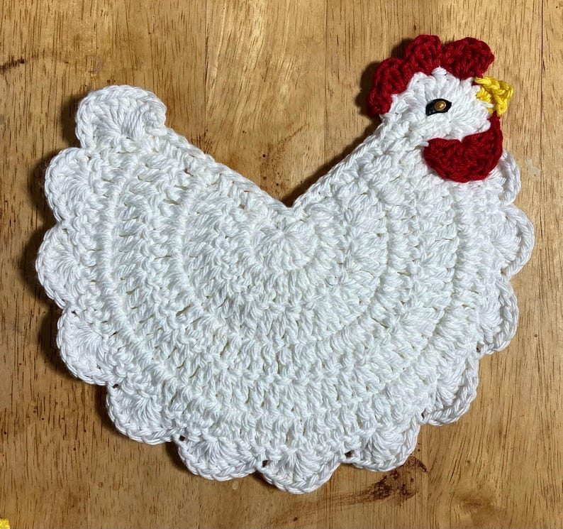 Pode incluir: Um porta-copos ou aplica&ccedil;&atilde;o de croch&ecirc; branco em forma de galinha com borda festonada. O design apresenta uma crista e barbelas vermelho-brilhantes, um pequeno bico amarelo e um olho escuro, tudo feito com pontos de l&atilde; detalhados.