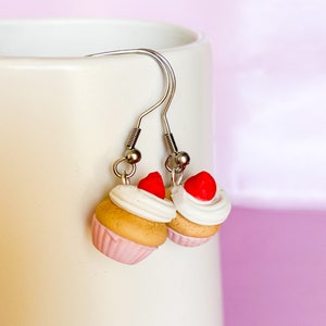 Puede incluir: Un par de pendientes de plata con colgantes de cupcake. Los cupcakes están decorados con glaseado blanco, fresas rojas y forros de cupcake rosas.