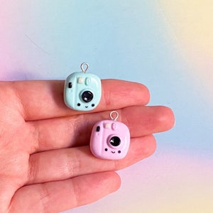 Puede incluir: Dos encantos de cámara en miniatura, uno azul claro y otro rosa, con detalles negros y una cara sonriente. Los encantos están en una mano.