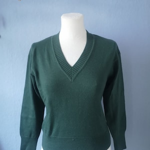 Könnte beinhalten: Ein dunkelgrüner V-Ausschnitt-Pullover, der auf einer Schaufensterpuppe präsentiert wird. Der Pullover hat lange Ärmel und Rippendetails am Ausschnitt, an den Manschetten und am Saum. Der Hintergrund ist eine hellblaue Wand mit einem dekorativen Ast.