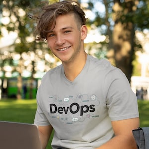 Peut inclure: Un t-shirt gris clair à col en V avec le mot "DevOps" en noir, ainsi que des icônes connexes. La personne est assise à l'extérieur avec un ordinateur portable et un sac à dos. L'arrière-plan est un parc avec des arbres et un bus.
