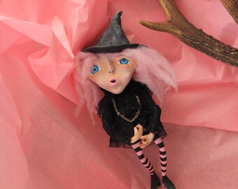 May include: A handmade witch doll with pink hair, blue eyes, and a black witch hat and dress. The doll has striped pink and black legs and a gold chain necklace. The doll is posed on pink tissue paper.