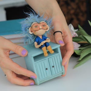 May include: A quirky figurine of a character with blue hair and a blue outfit, sitting on a small, pale blue cabinet. The figurine has a detailed face and is wearing tan boots. The cabinet has two doors and a small shelf.