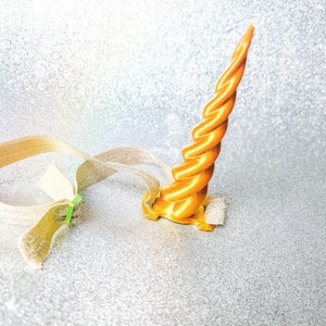 May include: A gold unicorn horn headband with a twisted design. The headband is made of a soft, flexible material and is attached to a brown elastic band.