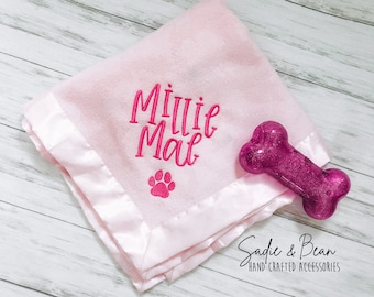 May include: Pink fleece blanket with a satin border. The blanket is embroidered with the name "Milli Mae" and a pink paw print. A pink bone toy is also pictured.