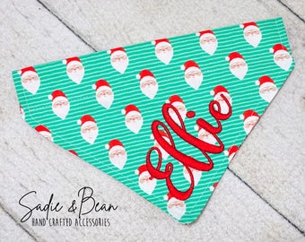May include: A turquoise and red dog bandana with a repeating pattern of Santa Claus faces. The bandana is personalized with the name "Ellie" in red embroidery.
