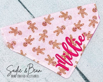 May include: Pink dog bandana with a gingerbread man pattern and the name "Millie" in pink lettering. The bandana is made by Sadie & Bean Hand-Crafted Accessories.