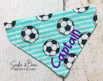 Peut inclure: Un bandana pour chien bleu turquoise à rayures blanches et des ballons de football noirs et blancs. Le bandana a le mot "Capitaine" brodé en violet.