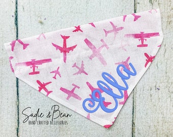 Peut inclure: Un bandana blanc avec un motif d'avion rose et le nom "Ella" brodé en bleu. Le bandana porte le texte "Sadie & Bean Hand-Crafted Accessories" en bas.