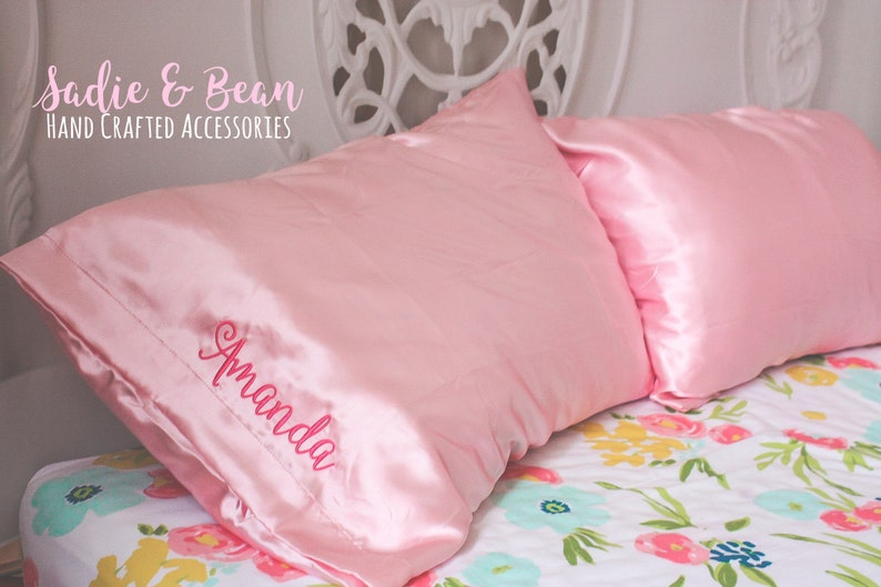 May include: Two pink satin pillowcases with the name "Amanda" embroidered on one of the pillowcases. The pillowcases are on a bed with a floral patterned bedspread.