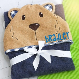 May include: A navy blue hooded baby towel with a bear face design. The towel features a tan bear face with blue gingham accents and the name "BRADLEY" embroidered in blue. A white ribbon wraps the towel.