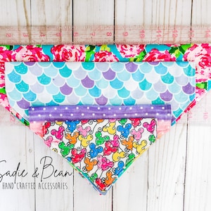 May include: Four different patterned dog bandanas stacked on top of each other. The bandanas are pink with flowers, blue with scales, purple with polka dots, and white with colorful balloons and Mickey Mouse heads. The bandanas are laid out on a white wooden surface with a ruler behind them. The text "Sadie & Bean HAND CRAFTED ACCESSORIES" is at the bottom of the image.