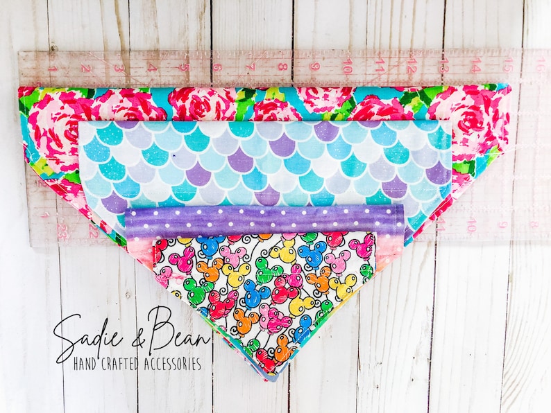 May include: Four different patterned dog bandanas stacked on top of each other. The bandanas are pink with flowers, blue with scales, purple with polka dots, and white with colorful balloons and Mickey Mouse heads. The bandanas are laid out on a white wooden surface with a ruler behind them. The text "Sadie & Bean HAND CRAFTED ACCESSORIES" is at the bottom of the image.