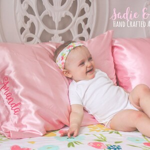 May include: A pink satin pillow with the name "Amanda" embroidered on it. A baby girl is lying on a floral bedspread with a pink satin pillow behind her head. She is wearing a white onesie and a floral headband. The text "Sadie & Bean HAND CRAFTED ACCESSORIES" is at the top of the image.