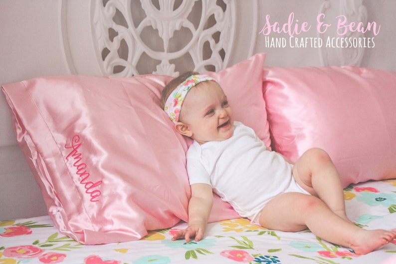 May include: A pink satin pillow with the name "Amanda" embroidered on it. A baby girl is lying on a floral bedspread with a pink satin pillow behind her head. She is wearing a white onesie and a floral headband. The text "Sadie & Bean HAND CRAFTED ACCESSORIES" is at the top of the image.