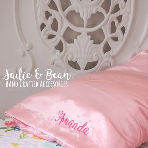 May include: A pink satin pillow with the name "Amanda" embroidered in pink on the front. The pillow is on a bed with a floral patterned sheet. The text "Sadie & Bean HAND CRAFTED ACCESSORIES" is visible in the top left corner of the image.