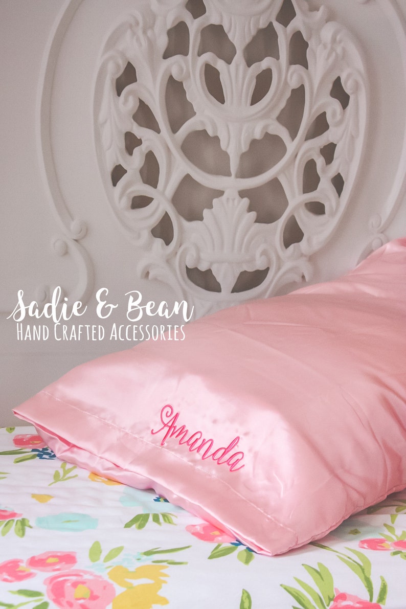 May include: A pink satin pillow with the name "Amanda" embroidered in pink on the front. The pillow is on a bed with a floral patterned sheet. The text "Sadie & Bean HAND CRAFTED ACCESSORIES" is visible in the top left corner of the image.
