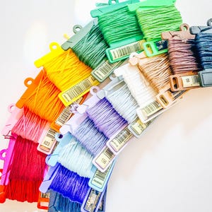 May include: A collection of embroidery floss skeins wrapped around plastic bobbins, arranged in a vibrant rainbow arc. The colors include red, pink, orange, yellow, green, light blue, purple, white, tan, brown, and dark blue, each with a barcode.