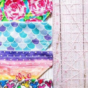 May include: Four colorful fabric dog bandanas with different patterns. The bandanas are lined up vertically with a pink ruler to the right. The bandanas feature floral, mermaid scale, polka dot, and Mickey Mouse balloon patterns. The top bandana has the text "Sadie & Bean" in pink.