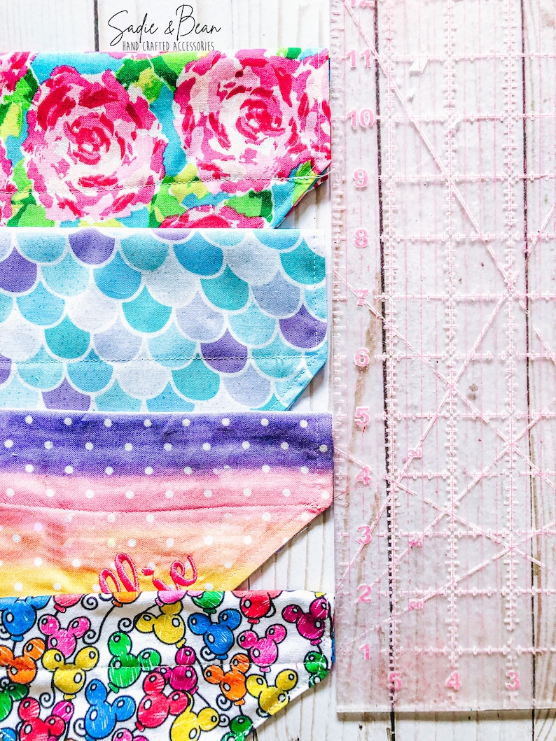 May include: Four colorful fabric dog bandanas with different patterns. The bandanas are lined up vertically with a pink ruler to the right. The bandanas feature floral, mermaid scale, polka dot, and Mickey Mouse balloon patterns. The top bandana has the text "Sadie & Bean" in pink.