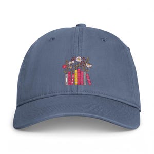 May include: A denim blue baseball cap featuring an embroidered graphic of a stack of colorful books in shades of pink, yellow, and grey, topped with various small white and red flowers and green stems centered on the front panel of the hat.
