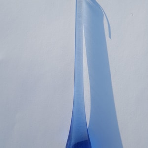 May include: A long, teardrop-shaped blue glass ornament with a gold-coloured hook at the top. The ornament casts a long shadow on a white surface. The glass has a slight texture.