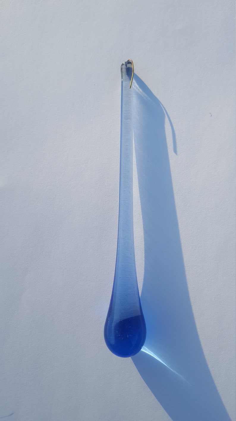 May include: A long, teardrop-shaped blue glass ornament with a gold-coloured hook at the top. The ornament casts a long shadow on a white surface. The glass has a slight texture.