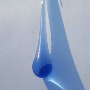 May include: A blue glass teardrop ornament with a silver loop at the top. The glass has a gradient from light to dark blue. The ornament's shadow stretches across a white surface.
