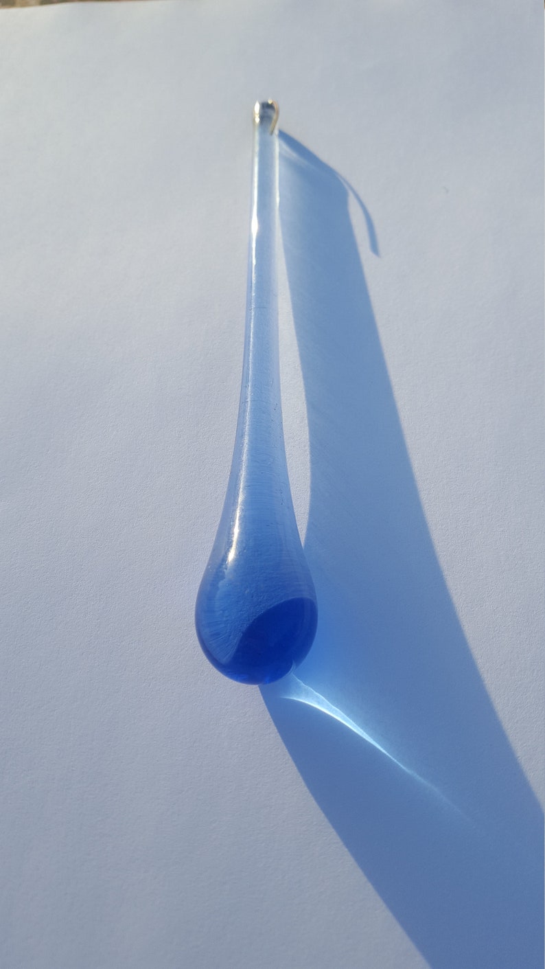 May include: A blue glass teardrop ornament with a silver loop at the top. The glass has a gradient from light to dark blue. The ornament's shadow stretches across a white surface.