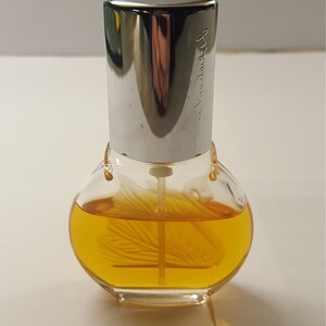 Op de afbeelding: Een heldere glazen parfumfles met een zilveren dop. De fles bevat een gele vloeistof en heeft een decoratief ontwerp aan de binnenkant. De dop is gegraveerd met de tekst "via Vandore".