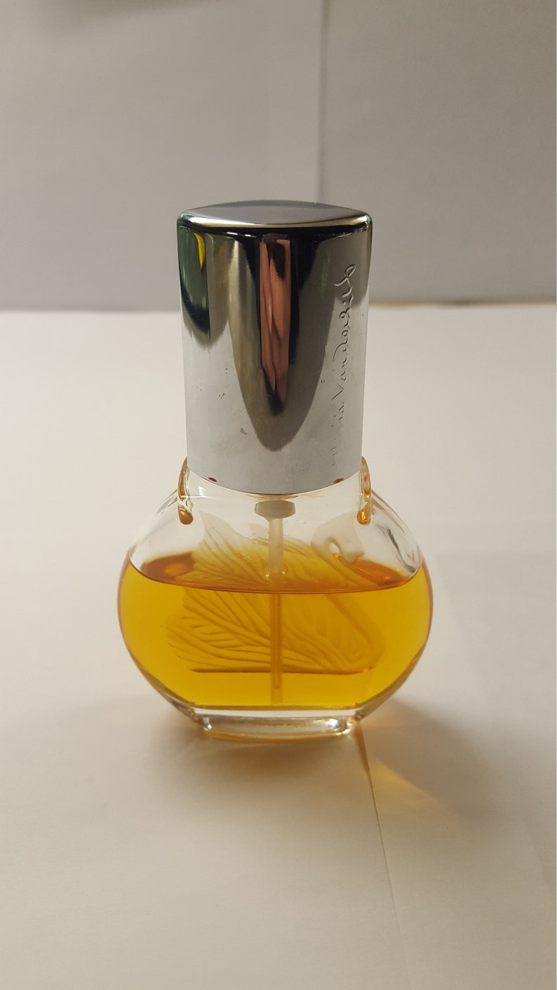 Op de afbeelding: Een heldere glazen parfumfles met een zilveren dop. De fles bevat een gele vloeistof en heeft een decoratief ontwerp aan de binnenkant. De dop is gegraveerd met de tekst "via Vandore".