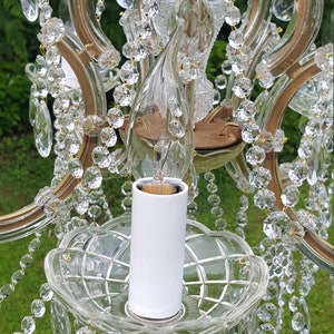 May include: Close-up of a vintage chandelier with clear crystal droplets and accents. The chandelier features a white candle holder and a glass light bulb. The frame is a gold color. The background is blurred green.