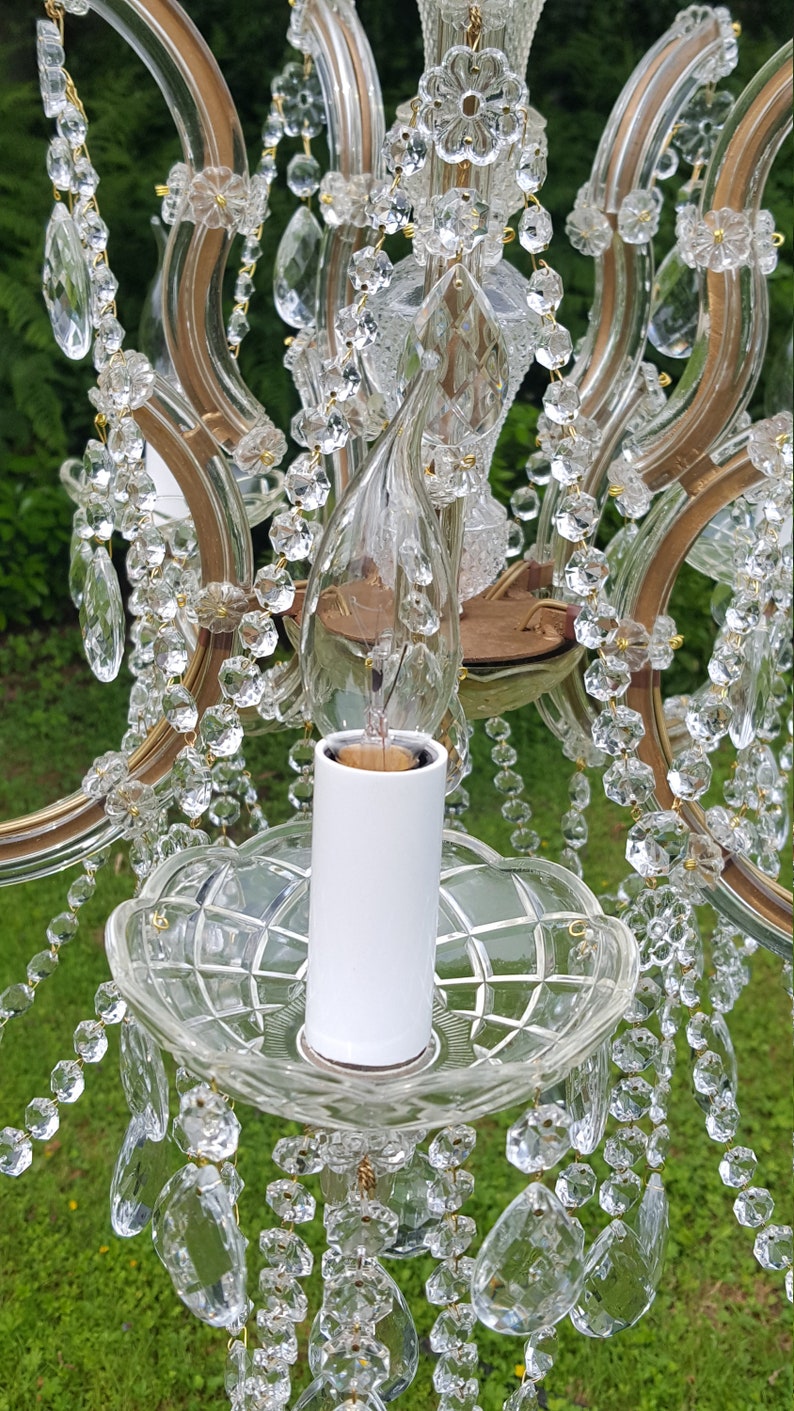 May include: Close-up of a vintage chandelier with clear crystal droplets and accents. The chandelier features a white candle holder and a glass light bulb. The frame is a gold color. The background is blurred green.