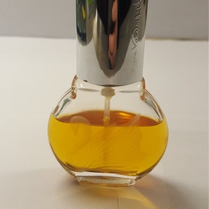 Op de afbeelding: Een kleine, heldere glazen parfumfles met een zilveren dop. De fles is rond met een zilveren dop en bevat een goudgele vloeistof. De naam "Mariah Carey" is op de zilveren dop ge&euml;tst. De fles staat op een wit oppervlak.