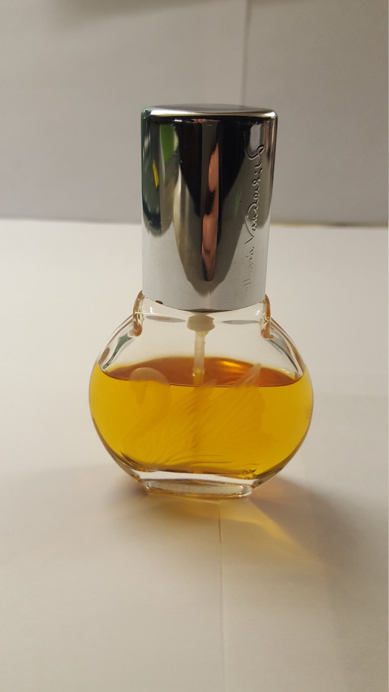 Op de afbeelding: Een kleine, heldere glazen parfumfles met een zilveren dop. De fles is rond met een zilveren dop en bevat een goudgele vloeistof. De naam "Mariah Carey" is op de zilveren dop ge&euml;tst. De fles staat op een wit oppervlak.