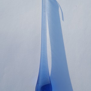 May include: A blue glass teardrop ornament with a gold-coloured hook. The teardrop shape is elongated, with a rounded bottom and a narrow top. The ornament casts a long shadow on a white surface.