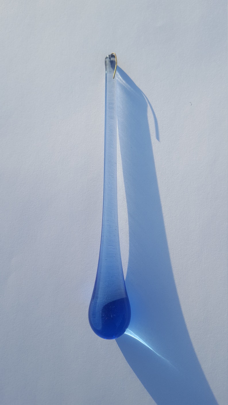 May include: A blue glass teardrop ornament with a gold-coloured hook. The teardrop shape is elongated, with a rounded bottom and a narrow top. The ornament casts a long shadow on a white surface.