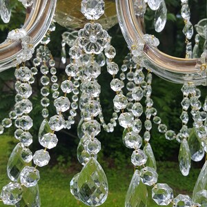 May include: Close-up of a crystal chandelier with multiple strands of clear, faceted crystals. The chandelier has a gold-colored frame and is set against a blurred green background. The crystals are various shapes and sizes.