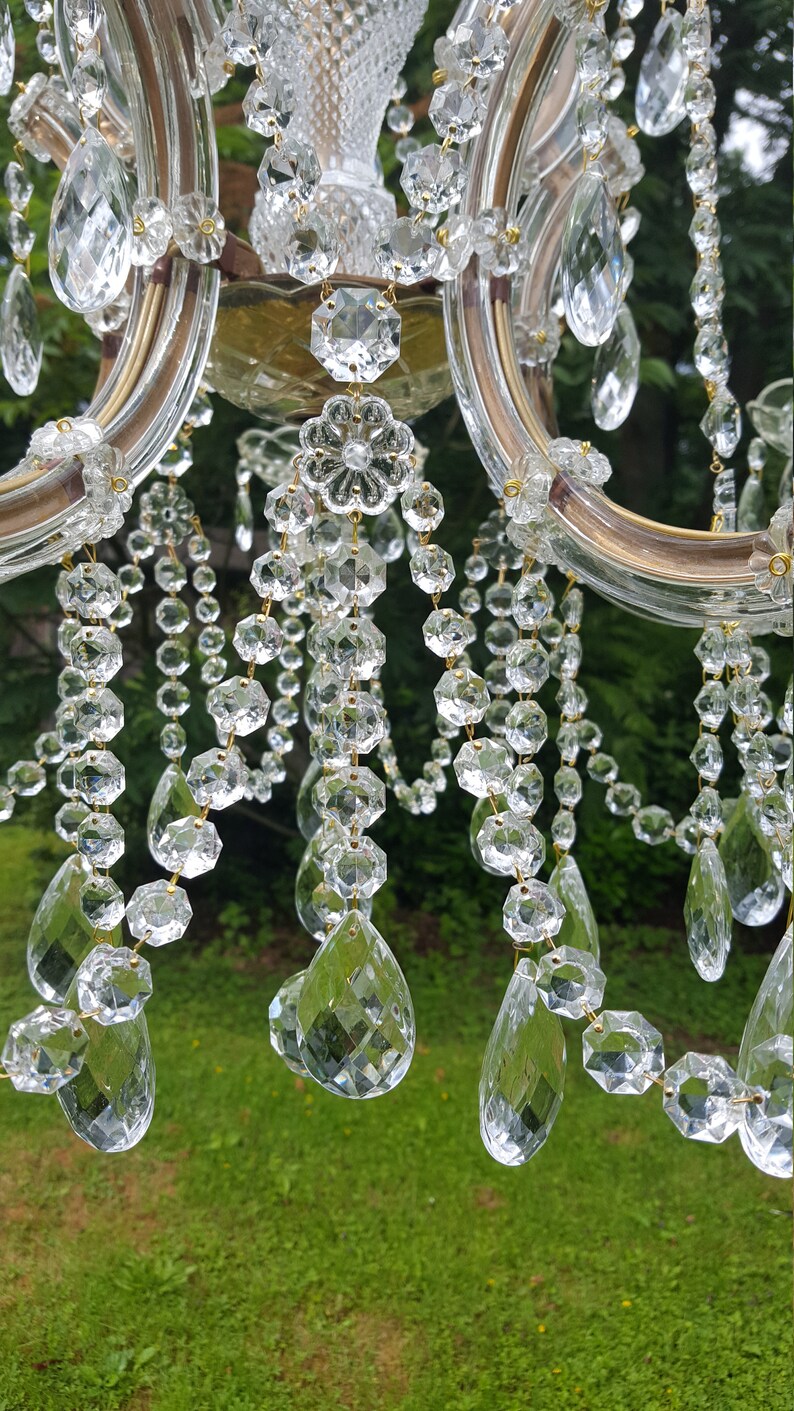 May include: Close-up of a crystal chandelier with multiple strands of clear, faceted crystals. The chandelier has a gold-colored frame and is set against a blurred green background. The crystals are various shapes and sizes.