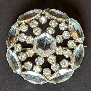 以下が含まれることがあります： 中央に大きな透明なファセットカットのクリスタルがついた、丸い金色のブローチ。クリスタルは、より小さな透明なファセットカットのクリスタルで囲まれています。