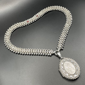 May include: A silver locket necklace with a detailed engraved design. The chain is made of a series of linked silver beads.