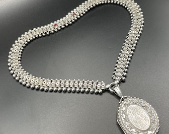 Pode incluir: Um colar de prata com um medalhão com um design gravado detalhado. A corrente é feita de uma série de contas de prata interligadas.