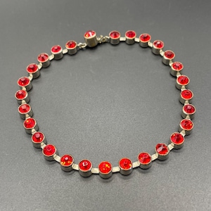 May include: A silver-toned necklace featuring a series of vibrant red gemstones. The round, faceted stones are set in individual bezels, creating a continuous loop. The necklace has a classic design, suitable for various occasions.