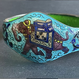 Puede incluir: Un brazalete de plata con un diseño de esmalte azul de un elefante que lleva una caja decorada. El elefante está contorneado en negro y tiene detalles morados, amarillos y blancos. El fondo es azul claro con remolinos blancos.