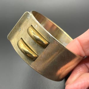 May include: A wide, gold-toned metal cuff bracelet. The bracelet has a textured surface and two gold-coloured rectangular accents. The bracelet is held by a hand, showcasing its design. A vintage jewellery piece.